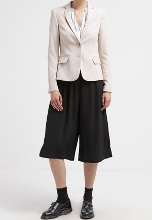 Vrouw die een beige blazer draagt over een gestreept overhemd, zwarte wijde culottes, zwarte sokken en zwarte leren nette schoenen.