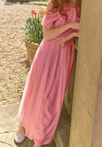 Lång rosa klänning i lätt tyg med avtagna ärmar, samlad midja och draperad kjol. Bärs med vita mönstrade skor.