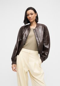 Brown leather bomber jacket with ribbed cuffs, front zipper, and a shiny finish, paired with a beige top and pleated cream trousers.