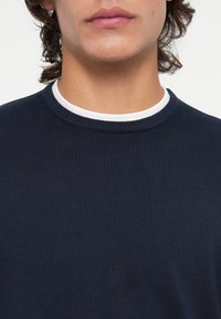 Pull bleu marine avec une texture côtelée, doté d'un col rond et d'un col intérieur blanc. Accent sur la zone de la poitrine sans motifs visibles.