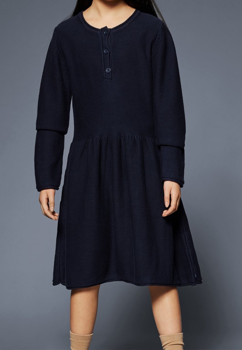 Enfant portant une robe tricotée bleu marine à manches longues avec des boutons au col et des chaussettes beiges, debout devant un fond gris.