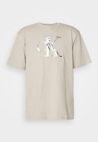 Beige cotton t-shirt with short sleeves and a round neckline. Features a large silver reflective "ck" logo on the front.