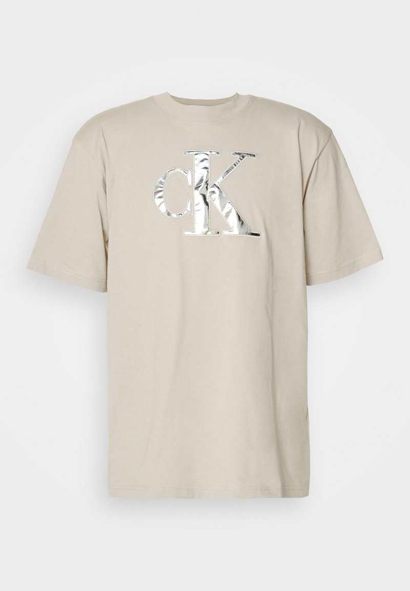 Beige cotton t-shirt with short sleeves and a round neckline. Features a large silver reflective "ck" logo on the front.