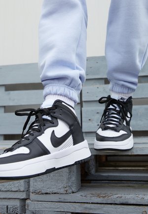 Osoba nosząca czarno-białe sneakersy Nike typu high-top oraz jasnoniebieskie spodnie dresowe, stojąca na szarych drewnianych paletach.