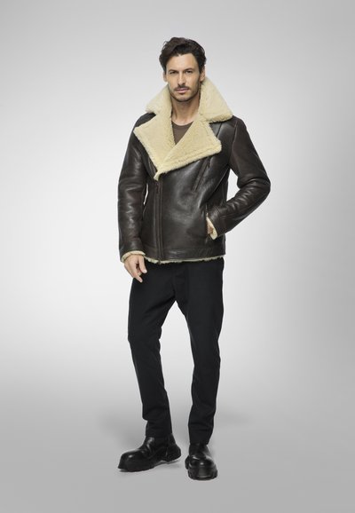 Giubbotto di pelle marrone con colletto in shearling crema, chiusura con zip e maniche lunghe. Abbinato a pantaloni neri e stivali neri robusti.