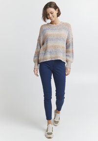 Strickpullover mitPastellstreifen in Beige, Blau und Orange, mit rundem Hals und lockeren Ärmeln, kombiniert mit blauen Jeans und hellen Turnschuhen.