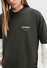 Camiseta de algodón de color verde oscuro, de manga corta y con cuello redondo. Presenta el logo en texto blanco "ALLSAINTS" impreso en el pecho.
