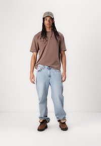 Brown cotton t-shirt, loose blue denim jeans, brown boots with tan accents, and a light beige cap. Casual outfit with relaxed fit.