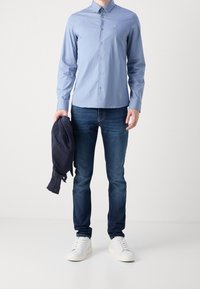 Uomo che indossa una camicia azzurro chiaro, jeans blu scuro, sneaker bianche, mentre tiene una giacca blu scuro nella mano sinistra, su uno sfondo neutro.
