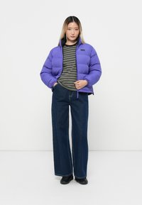 Giacca a vento viola con chiusura zip, polsini a coste e logo sul lato sinistro del petto, indossata sopra una maglietta a righe e abbinata a jeans larghi.