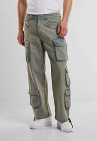 Cargo hlače u izblijedjelom plavom denimu s više bočnih džepova, slobodnog kroja i kontrastnim šavovima, uparene s bijelim tenisicama.