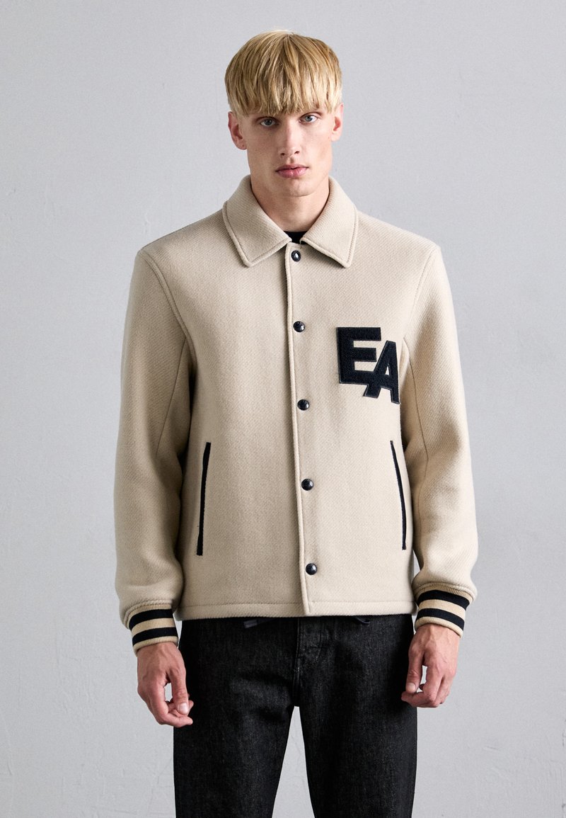 Μπεζ ζακέτα από μείγμα μάλλινου υφάσματος με κουμπιά μπροστά, μαύρες λεπτομέρειες, πλάγιες τσέπες και λογότυπο "EA". Διαθέτει ανοιχτό γιακά και ριμπ μανσέτες.