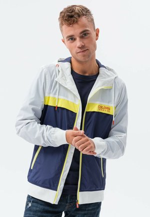 Heren zip-up jack in lichtgrijs, marineblauw en geel. Heeft een capuchon, elastische manchetten en een logo op de borst. Vloeiende textuur, lichtgewicht ontwerp.