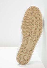 Semelle de chaussure avec une bande de roulement en caoutchouc beige présentant des motifs circulaires et de petites protubérances, appuyée contre un mur et un sol blancs.