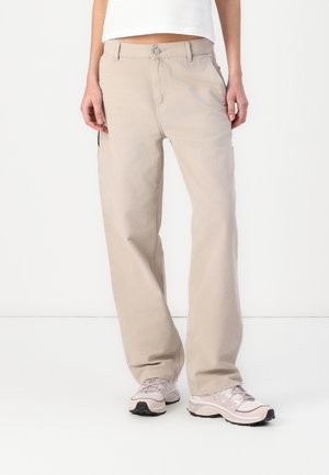 Person iført beige bukser med vide ben, hvid kort top og lyserøde sneakers, stående mod en ensfarvet hvid baggrund.