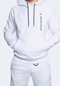 Sudadera blanca con capucha hecha de material suave con cordones oscuros, que presenta el logo "Calvin Klein Jeans" impreso verticalmente en negro.