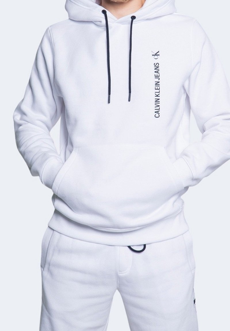Sudadera blanca con capucha hecha de material suave con cordones oscuros, que presenta el logo "Calvin Klein Jeans" impreso verticalmente en negro.