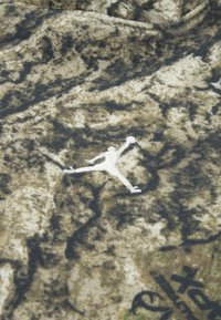 Tričko z látky s maskáčovým vzorom v zemitých odtieňoch hnedého a zeleného, s bielym logom Jumpman, ktoré je výrazne umiestnené.