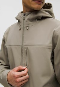 Chaqueta de softshell beige con cuello alto, cierre de cremallera y capucha ajustable. Presenta un bolsillo en el pecho con cremallera y textura suave.