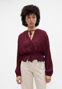 Blusa burgundy plissada com mangas longas, decote com laço e barra rendada. Usada com calças de cor clara, exibindo um tecido texturado.