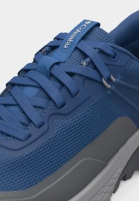 Κοντινό πλάνο μπλε αθλητικού παπουτσιού Columbia με υφαντό πλέγμα στην επάνω πλευρά, χοντρά μπλε κορδόνια και γκρι ελαστική σόλα.