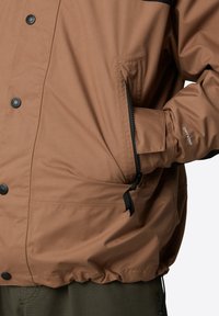 Brązowa wodoodporna kurtka z czarnymi akcentami, z zatrzaskami, zasuwanymi kieszeniami i elastycznym brzegiem dla regulowanego dopasowania.