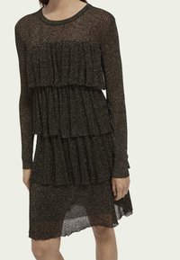 Schwarzes Glitzerkleid mit langen Ärmeln, gestuften Rüschen am Oberteil und Rock sowie einer leicht durchsichtigen Textur. Der Saum fällt hinten ab.