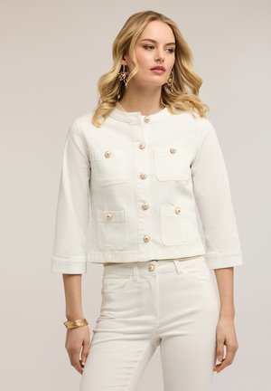 Blonde vrouw die een wit jasje draagt met vier zakken aan de voorkant, dichtgeknopt, en een bijpassende witte broek, gecombineerd met gouden oorbellen en een armband.