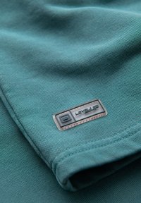 Nahaufnahme eines türkisfarbenen Stoffes mit einem gefalteten Eck und einem aufgenähten rechteckigen Abzeichen mit der Aufschrift "LEVEL UP" in Schwarz und Weiß.