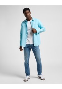 Camisa de botones azul claro con mangas largas, camiseta blanca debajo, jeans azul claro y zapatos deportivos en blanco y negro.
