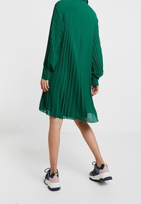 Vihreä laskostettu mekko pitkillä hihoilla, takana vetskari, ja rento leikkaus. Yhdistetty monivärisiin lenkkareihin, joissa on paksu pohja.