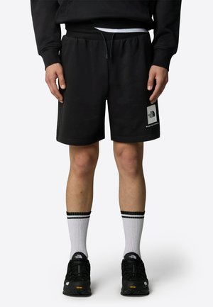 Sorte bomulds-shorts med elastisk talje, sportslogo-patch på benet, parret med sorte sko og hvide sokker med sorte striber.