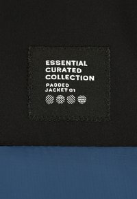 Padded-Jackenlabel mit schwarzem Stoff und weißem Text, der "Essential Curated Collection Padded Jacket 01" liest, sowie dekorativen Mustern.