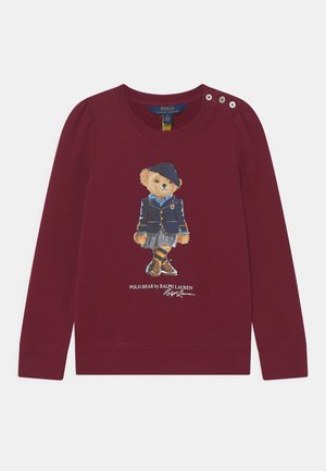 Viininpunainen pitkähihaispaita, jossa napit olkapäällä. Paidassa on kuvio karhusta, joka pukeutunut bleiseriin, hameeseen, baskeriin, raidallisiin sukkiin ja kenkiin.