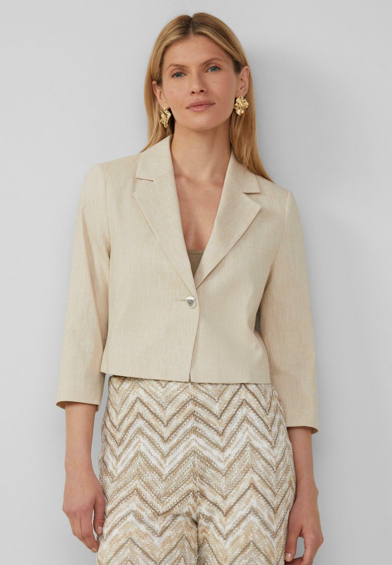 Beige cropped blazer met driekwart mouwen, met opgestikte kleppen en een enkele knoopsluiting, gecombineerd met beige broek met een patroon.