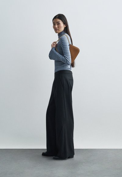 Donna in maglione a righe blu e pantaloni larghi neri, con una borsa marrone a tracolla con catena dorata appoggiata sulla spalla, contro un muro semplice.