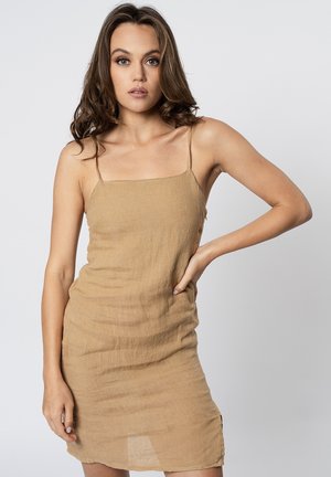 Giovane donna con capelli castani mossi che indossa un vestito senza maniche in lino color sabbia, in piedi con una mano sul fianco davanti a uno sfondo semplice.
