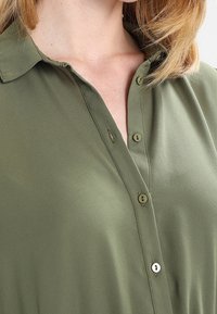 Oliwkowa bluzka o gładkiej fakturze, z zapięciem na guziki z przodu i klasycznym krojem kołnierza. Posiada małe guziki na listwie.