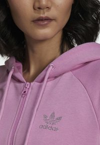 Rozā rāvējslēdzēja kapuce ar teksturētu audumu, kas rotāta ar sudraba rhinestone logos uz krūtīm un atbilstošiem auklām uz kapuces.