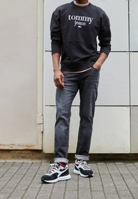 Uomo che indossa un felpa nera di Tommy Jeans, jeans grigio scuro con risvolti, calzini bordeaux e sneakers bianco e nere, in piedi su un terreno pavimentato.