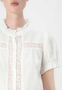Bijela bluza s kratkim, nabranim rukavima, teksturiranom tkaninom, čipkastim detaljima na tijelu i zatvaranjem na gumbe sprijeda.