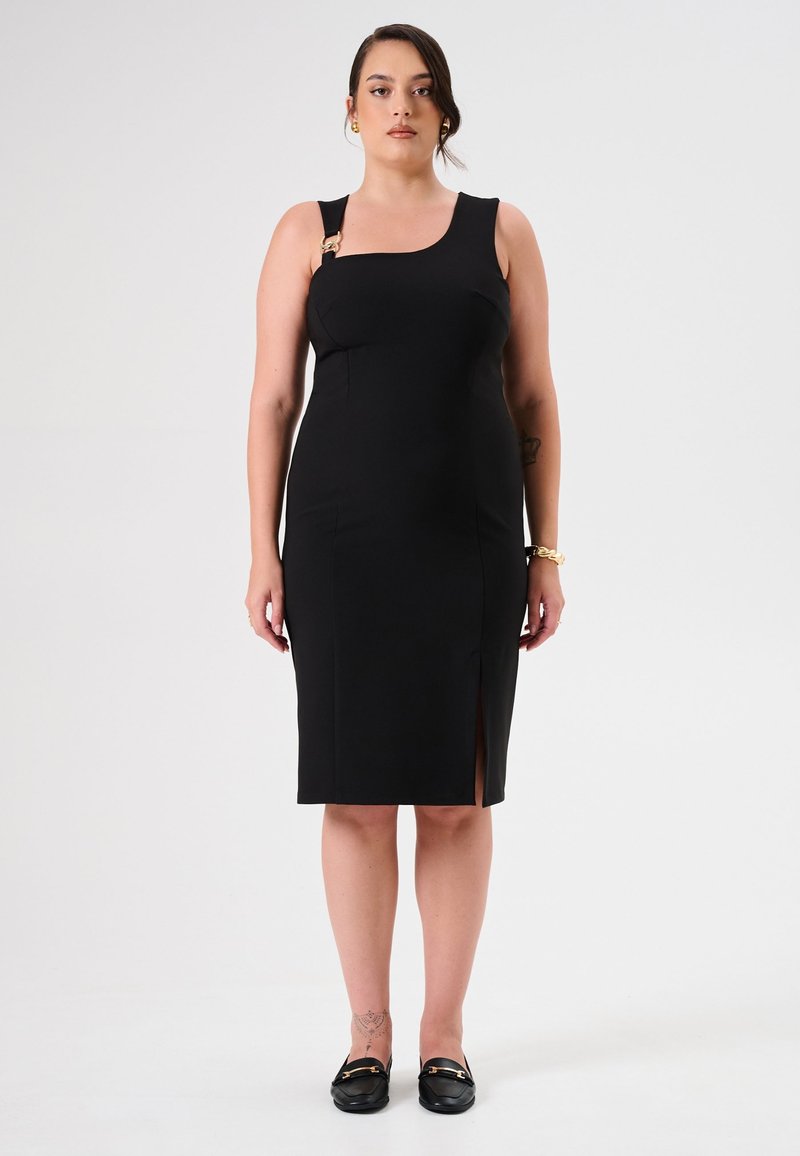 Schwarzes, figurbetontes Kleid mit quadratischem Ausschnitt, einem goldenen Akzent am Träger, seitlichem Schlitz und glatter Textur. Getragen mit schwarzen Slippern.