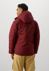 Maroon insulated jacket with a hood, featuring a smooth texture, elastic cuffs, and a slightly cinched waist, paired with beige cargo shorts.
