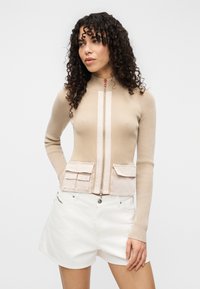 Μπεζ καζάκα με φερμουάρ και πλεκτή υφή με ρίγες, διαθέτει δύο τσέπες μπροστά και ψηλό γιακά. Συνδυάζεται με λευκά σορτς.