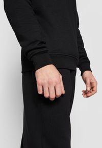 Man wearing black long-sleeve sweatshirt and black pants with hands relaxed at sides against plain background.