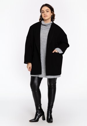 Sort oversized frakke over en grå ribbet sweaterkjole, parret med sorte læderleggings og ankelstøvler med strukturerede hæle.