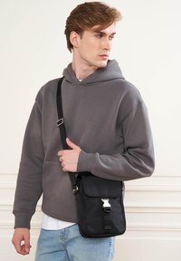 Borsa a tracolla nera con dettagli in argento, forma rettangolare, materiale testurizzato, indossata da una persona con una felpa grigia e jeans azzurri chiari.