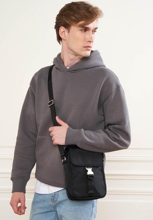 Schwarze Umhängetasche mit silbernen Details, rechteckige Form, strukturiertes Material, getragen von einer Person in einem grauen Hoodie und hellblauen Jeans.