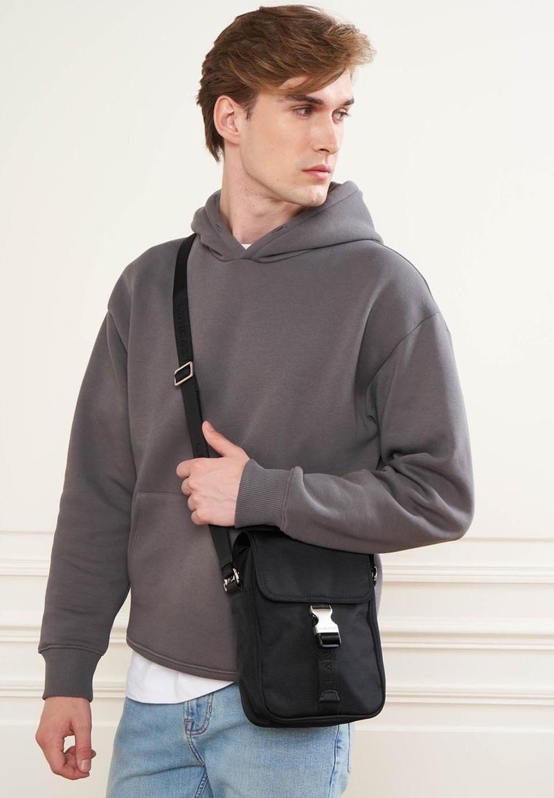 Borsa a tracolla nera con dettagli in argento, forma rettangolare, materiale testurizzato, indossata da una persona con una felpa grigia e jeans azzurri chiari.