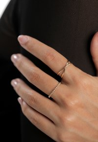 Twee gouden ringen aan een hand: één met een kruisontwerp en één met gelijkmatig verdeelde kralen. De hand rust tegen een getextureerd zwart oppervlak.